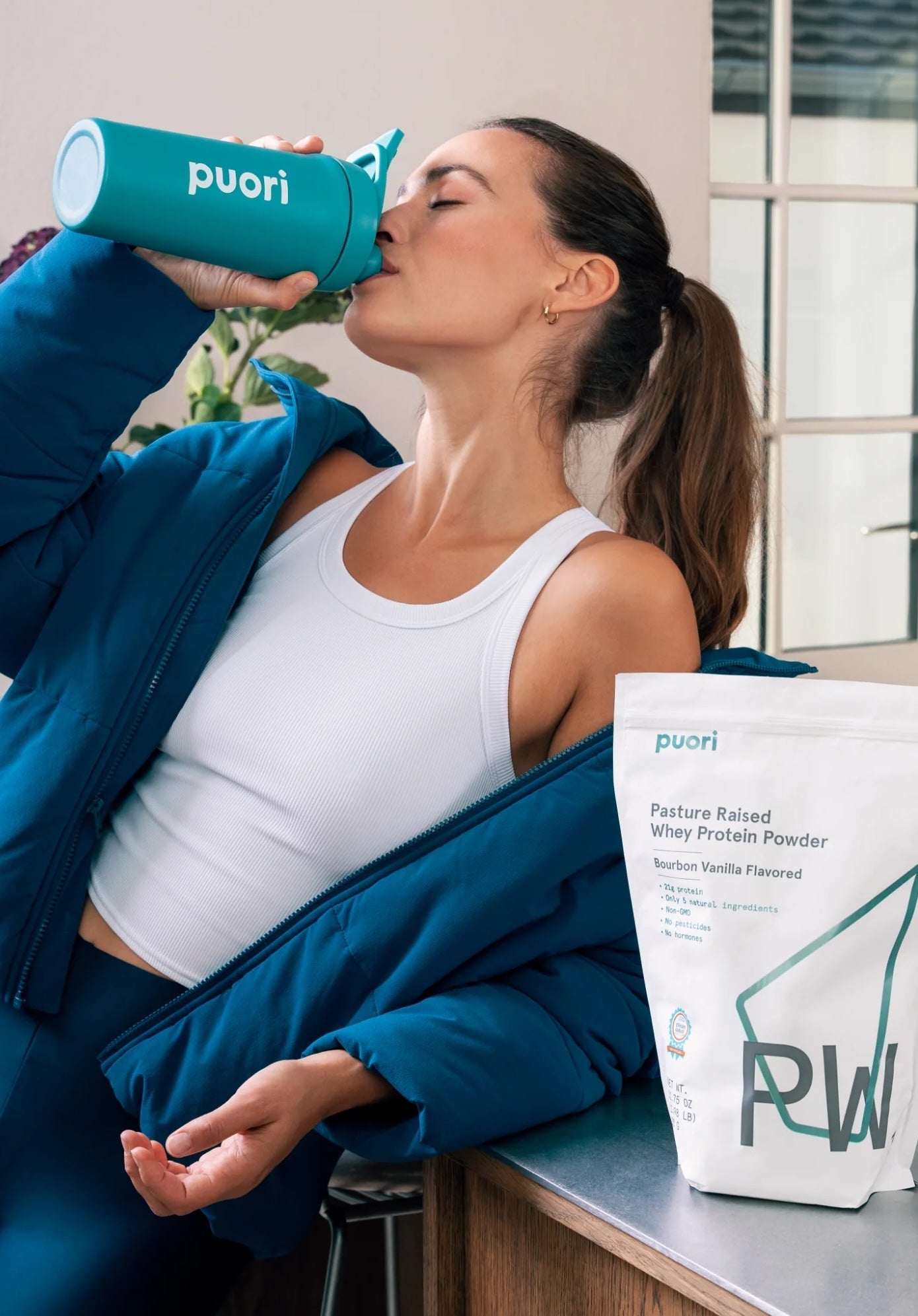 Woman in workout attire drinking from a teal Puori shaker bottle, with a Puori Pasture Raised Whey Protein Powder (Bourbon Vanilla) bag visible on the counter beside her.